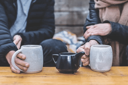 coffee vs. tea. A man and a woman sitting together holding onto gray mugs with a black kettle on the table between them